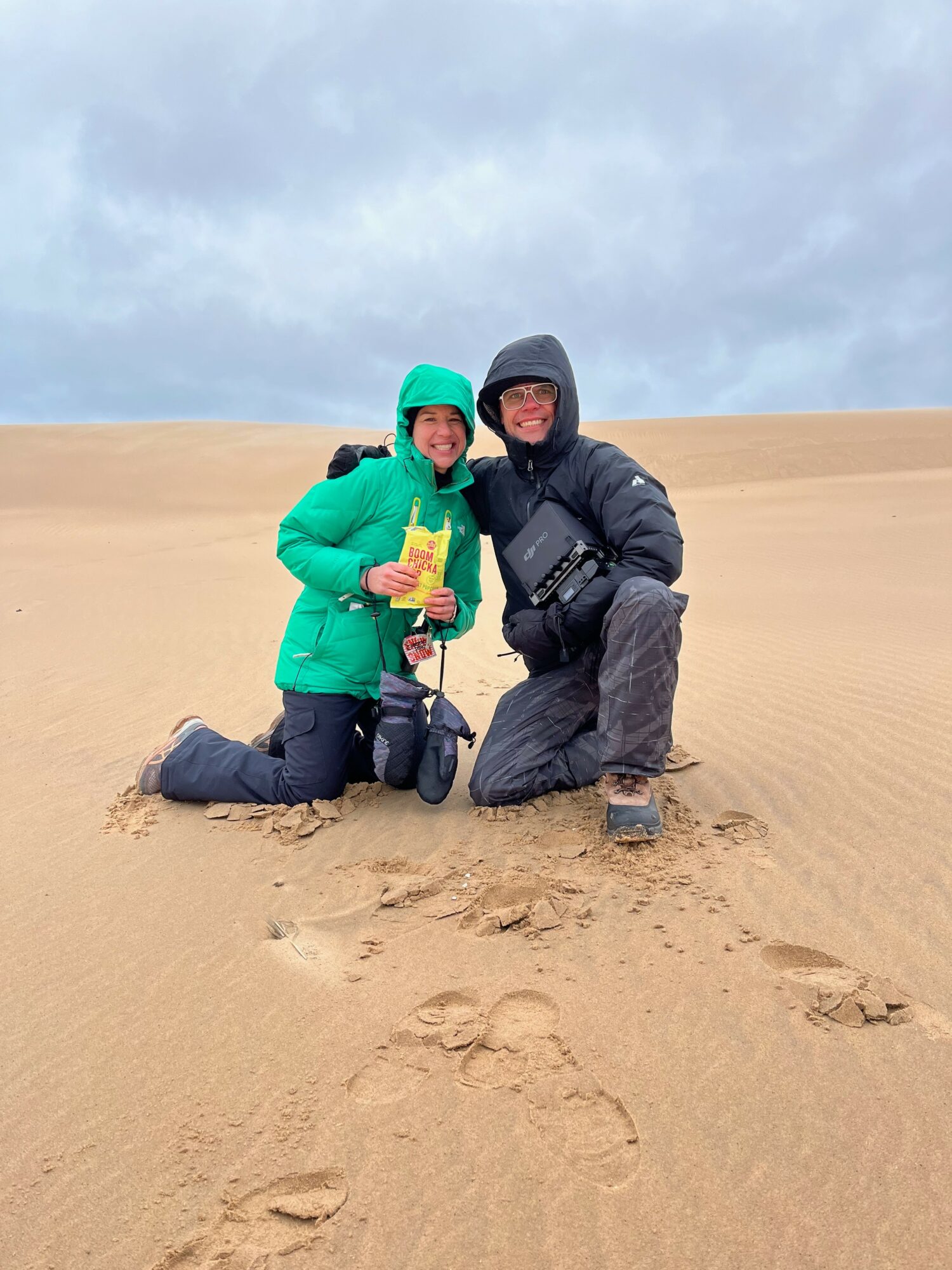 Two people kneeling on sand dunes, smiling, wearing jackets and hats, with footprints in the sand, cloudy sky above.