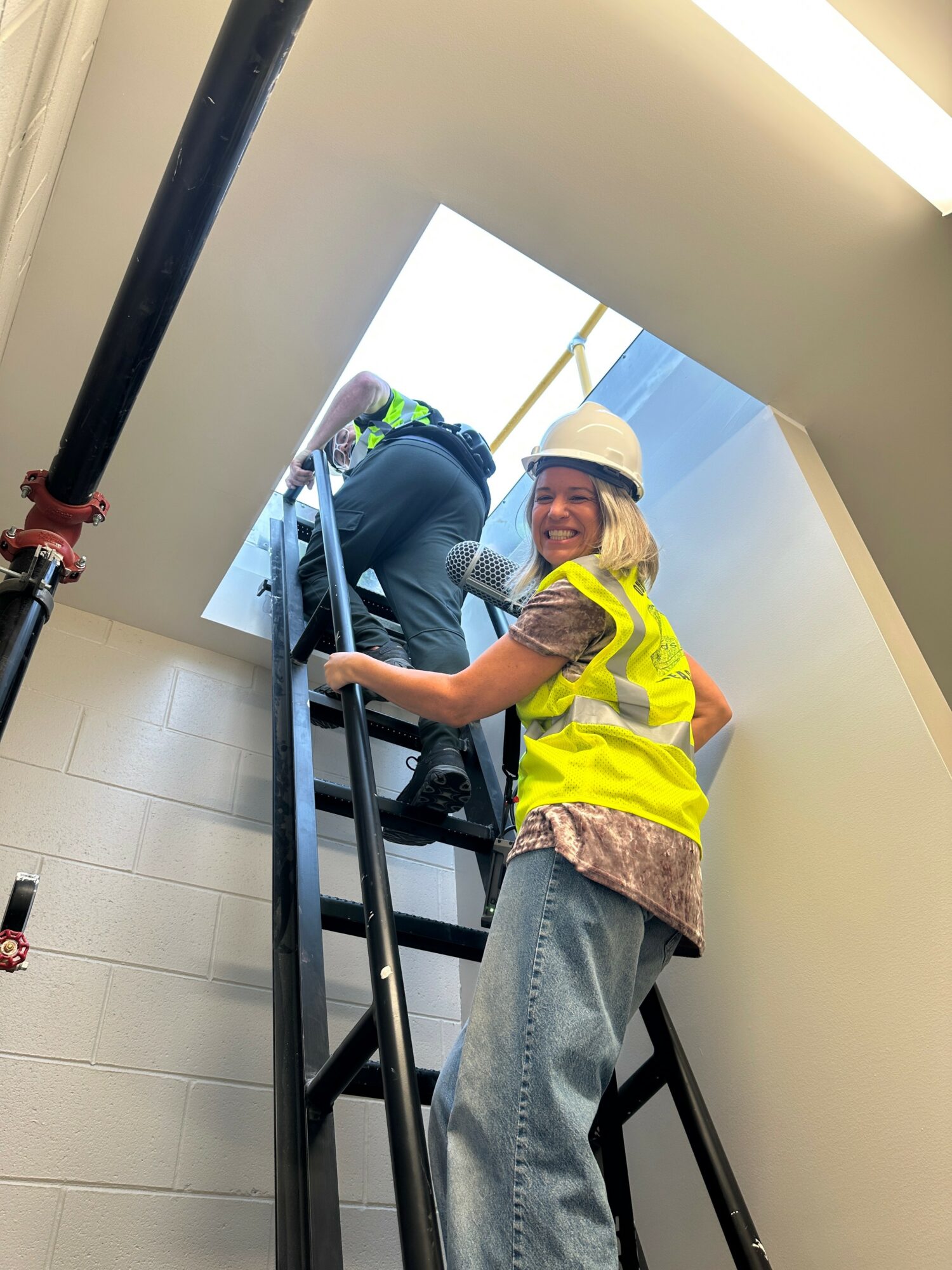 Woman in safety vest and helmet climbing stairs with a person at the top, inside a building.