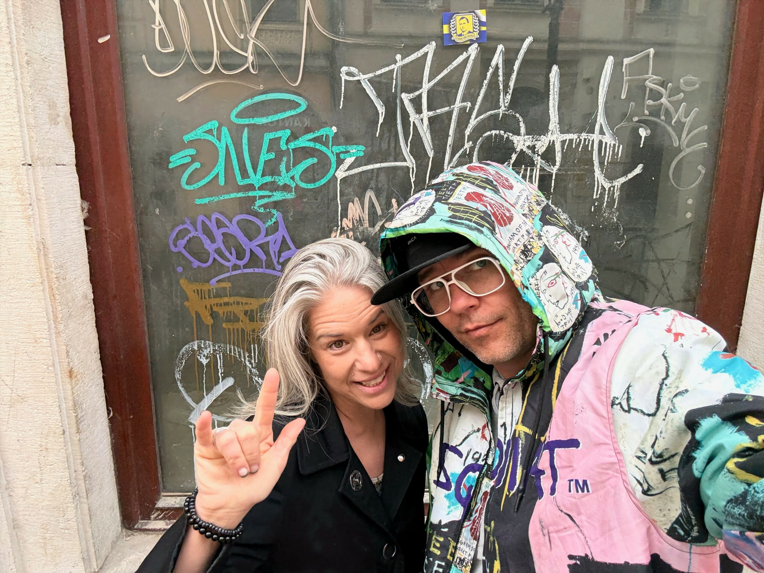 Two people taking a selfie in front of a graffiti-covered window, one with gray hair and the other wearing a colorful hoodie and glasses.