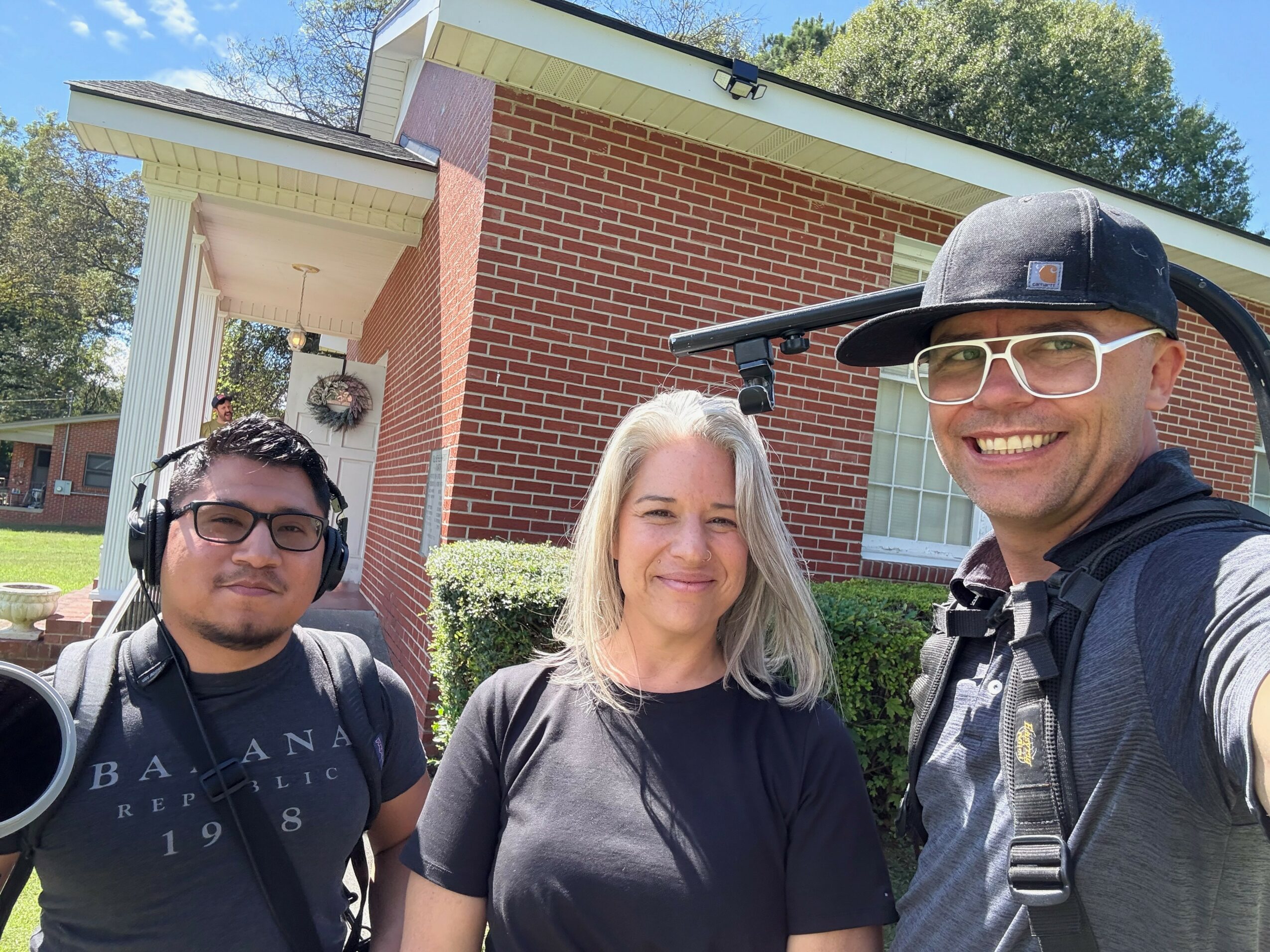 Three people smiling outdoors in front of a brick building, one taking a selfie, with trees in the background.