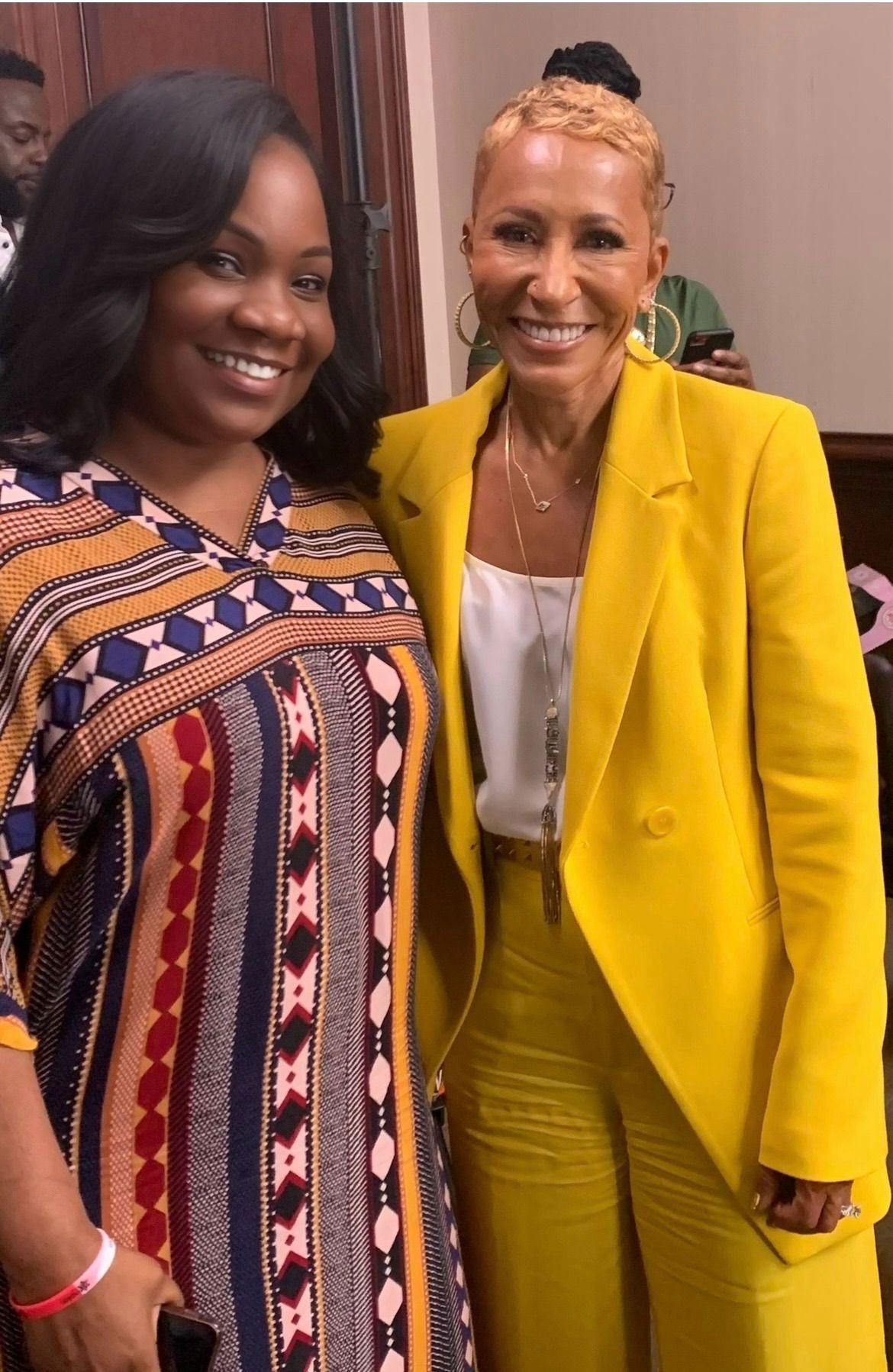 Two women smiling, one in a colorful patterned dress, the other in a yellow suit, standing indoors.