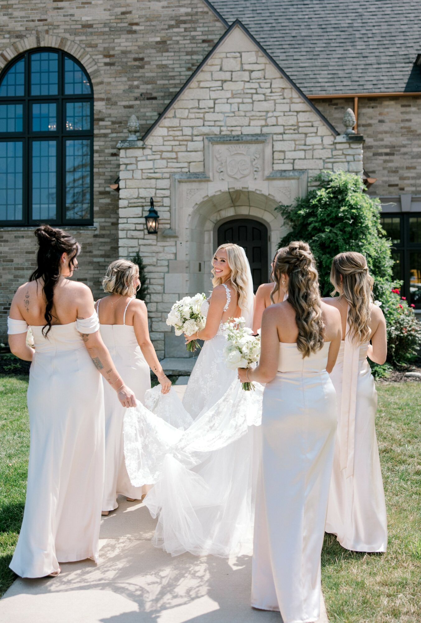 Bride in white dress with five bridesmaids in light-colored dresses outside a stone building, holding bouquets.
