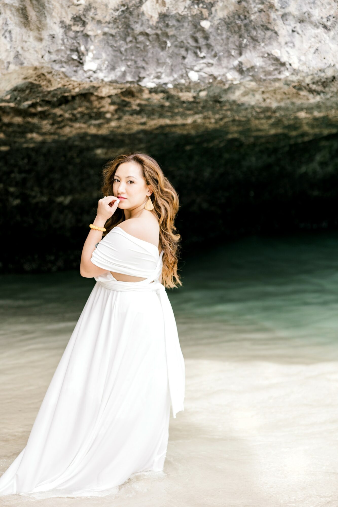 Woman in a white dress standing on a beach with a rocky overhang above her, facing sideways and looking at the camera.