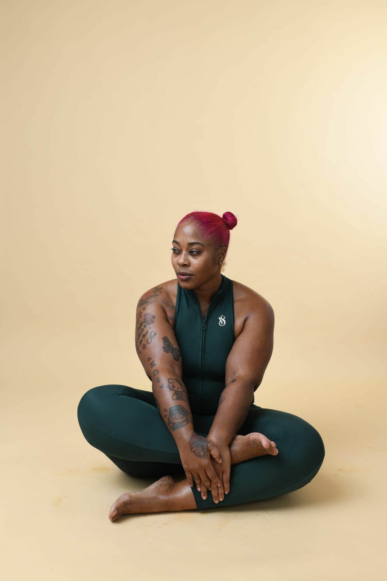 Person sitting cross-legged on the floor with a pink hair bun, wearing a dark sleeveless top, against a plain beige background.