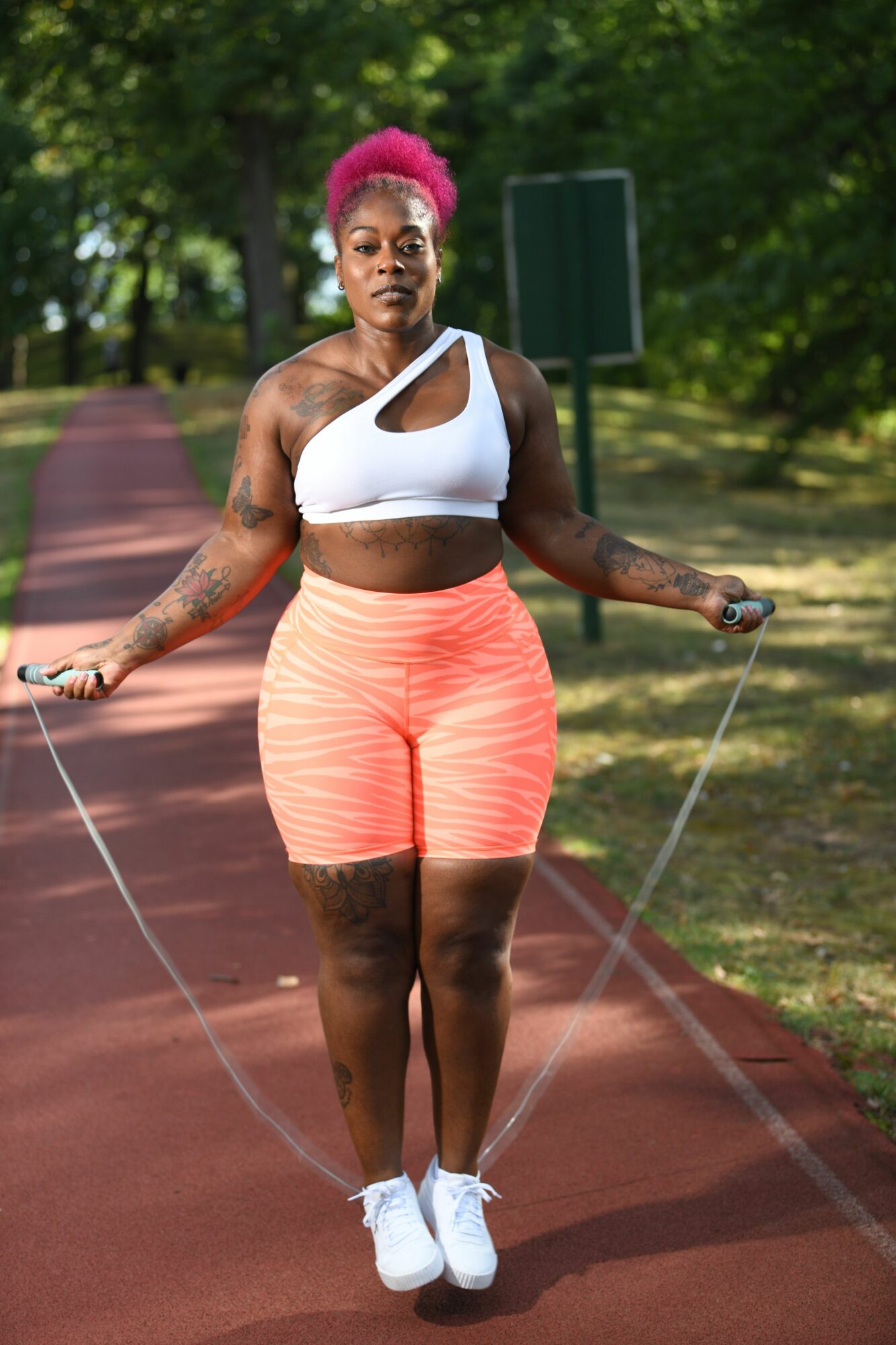 Woman with pink hair skipping rope on outdoor track, wearing athletic clothing, surrounded by trees.