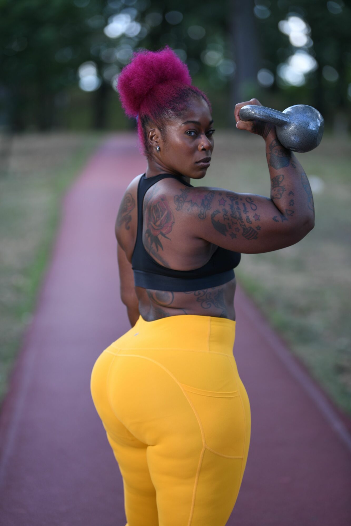 Woman with pink hair in a ponytail, wearing yellow pants and a black sports bra, lifting a dumbbell outdoors.