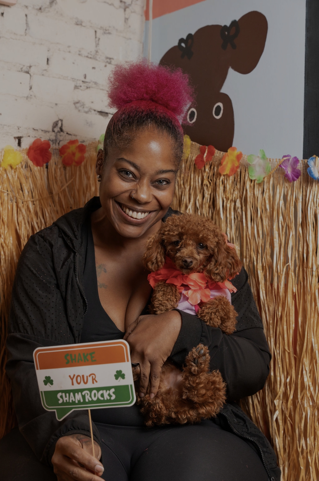 Woman with pink hair holding a small brown dog, smiling, in a decorated indoor setting with a sign reading 'Your Shamrocks'.