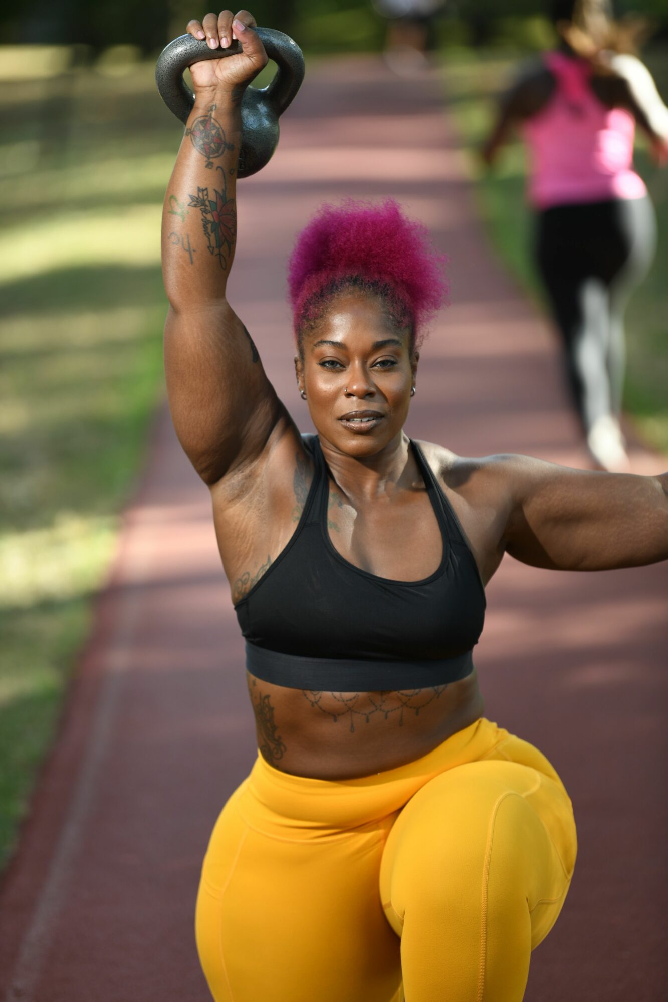 Woman with pink hair lifting a kettlebell outdoors on a track, wearing black sports bra and yellow pants.