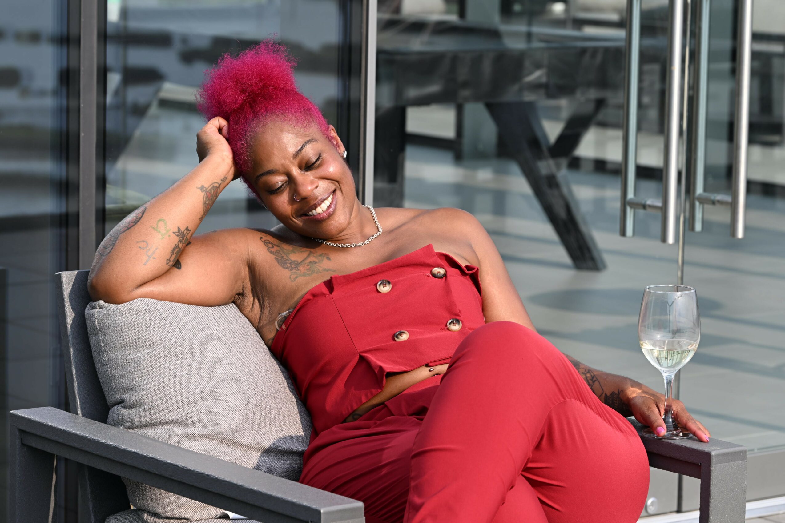 Woman with pink hair relaxing on a gray outdoor lounge chair, wearing a red strapless outfit, with a glass of water nearby.