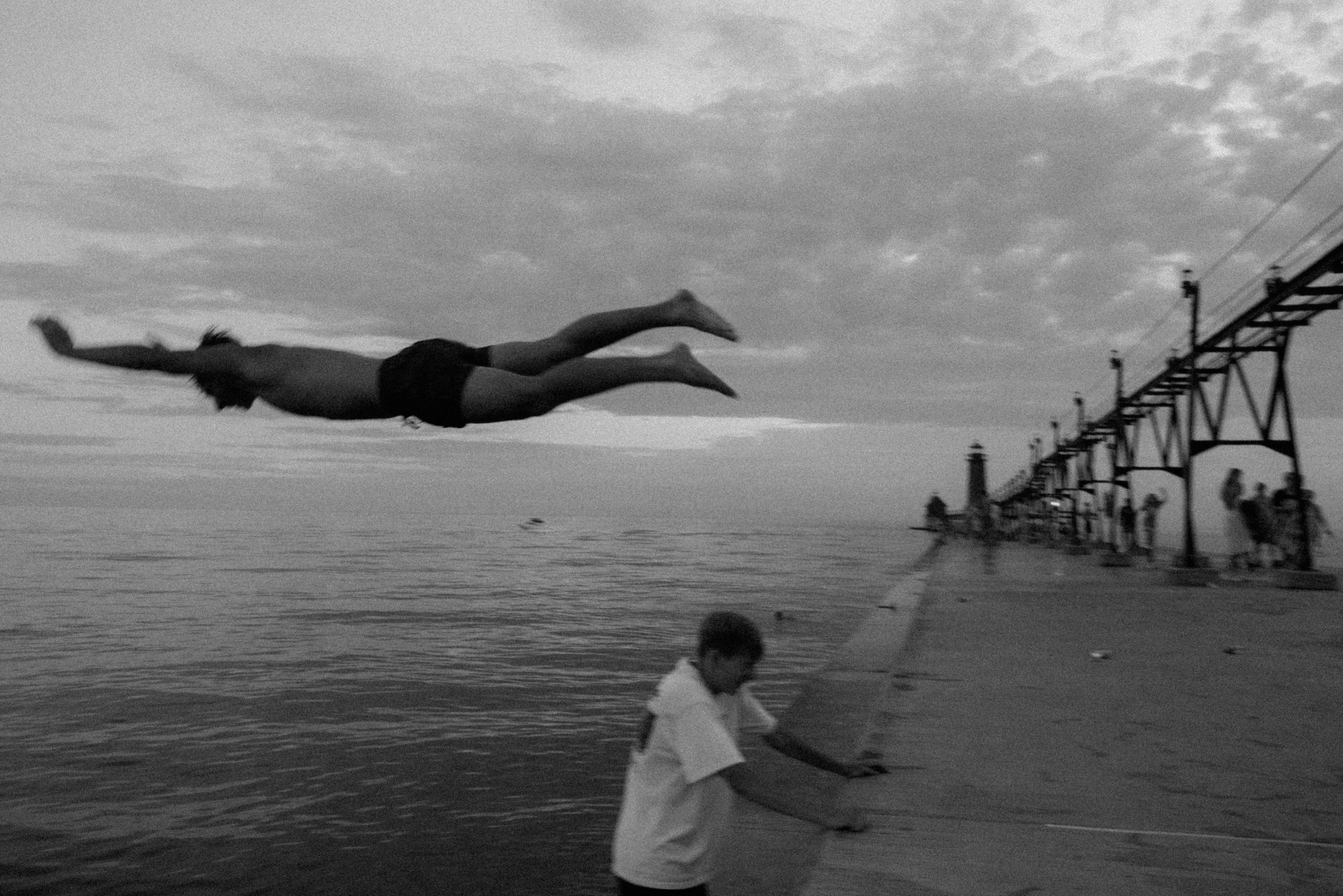 Person diving into water near a pier, with cloudy sky and calm sea in background.