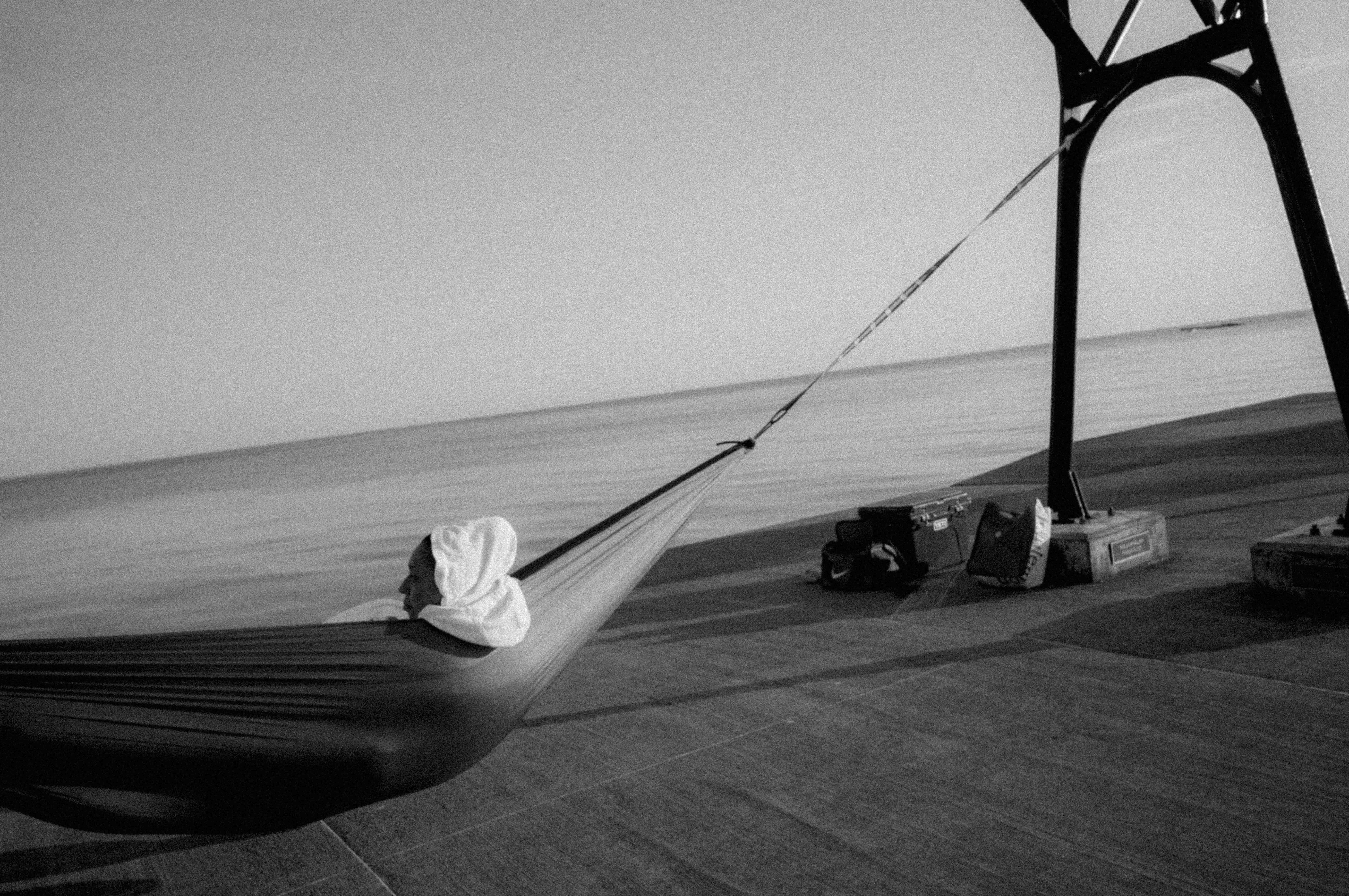 A hammock with a pillow is on a concrete surface near the water, with a boat lift and water in the background.