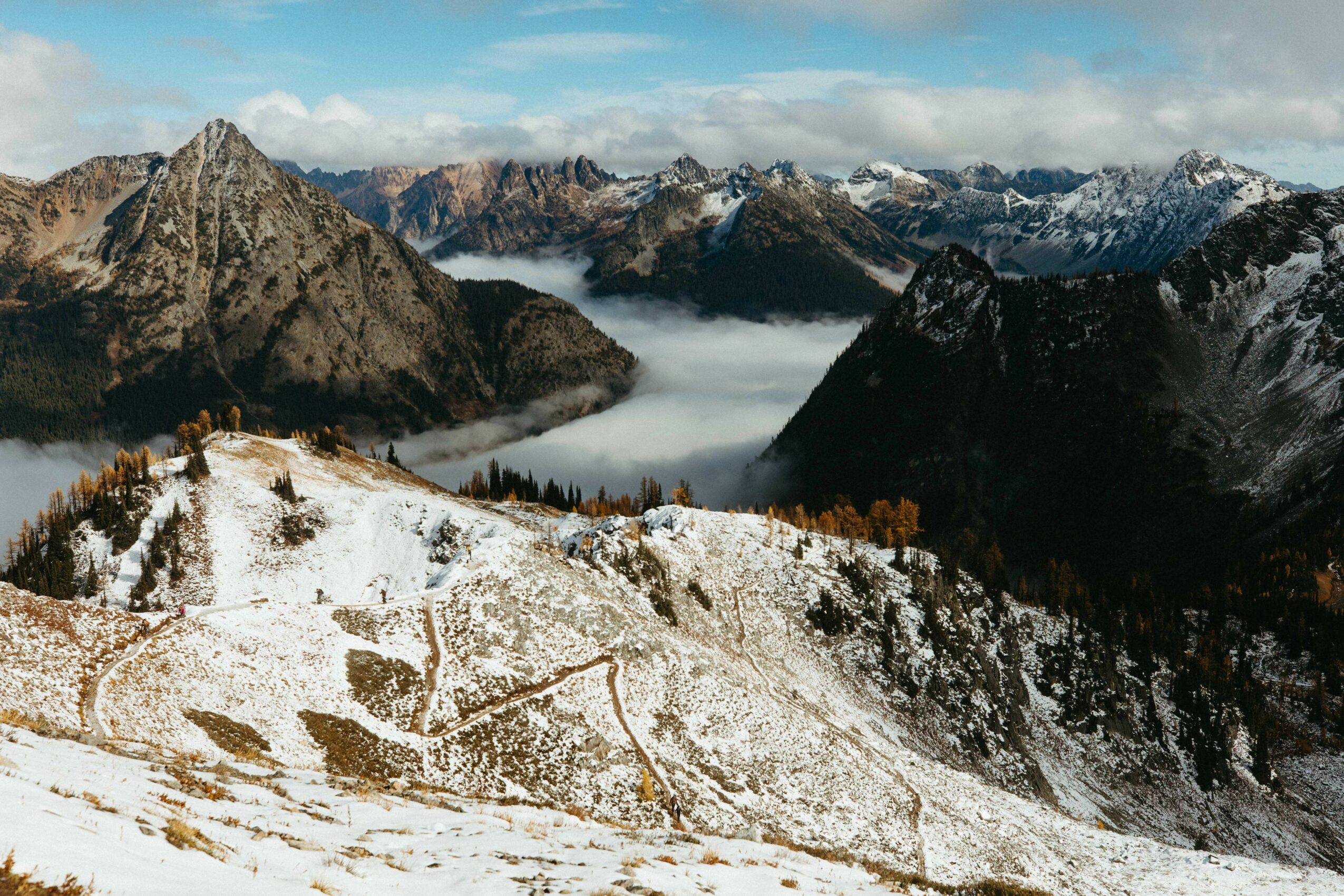 Snow-covered mountain landscape with rugged peaks, clouds, and fog in the valleys, under a partly cloudy sky.