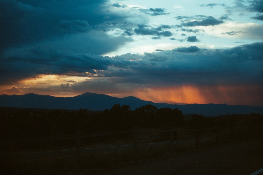 Cloudy sky with sunset, dark mountains in the distance, and a silhouette of a landscape in the foreground.