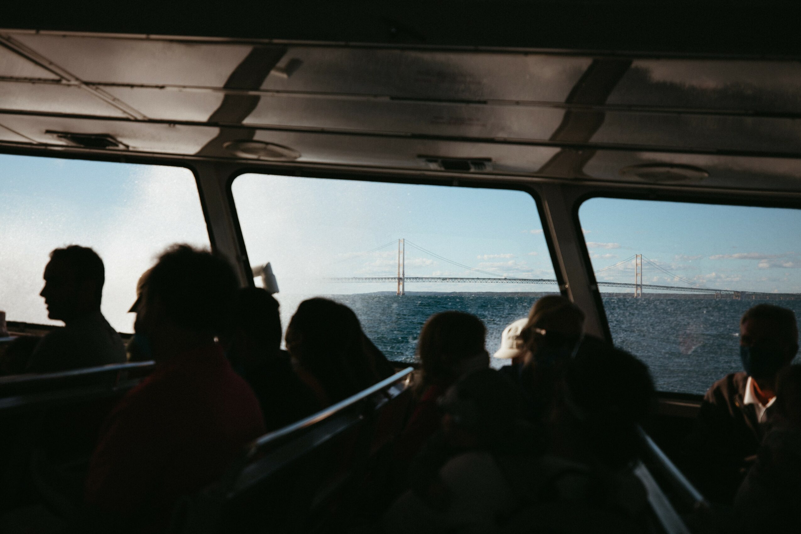 People sitting inside a boat or ferry, looking out the windows at a bridge and water, with a cloudy sky.
