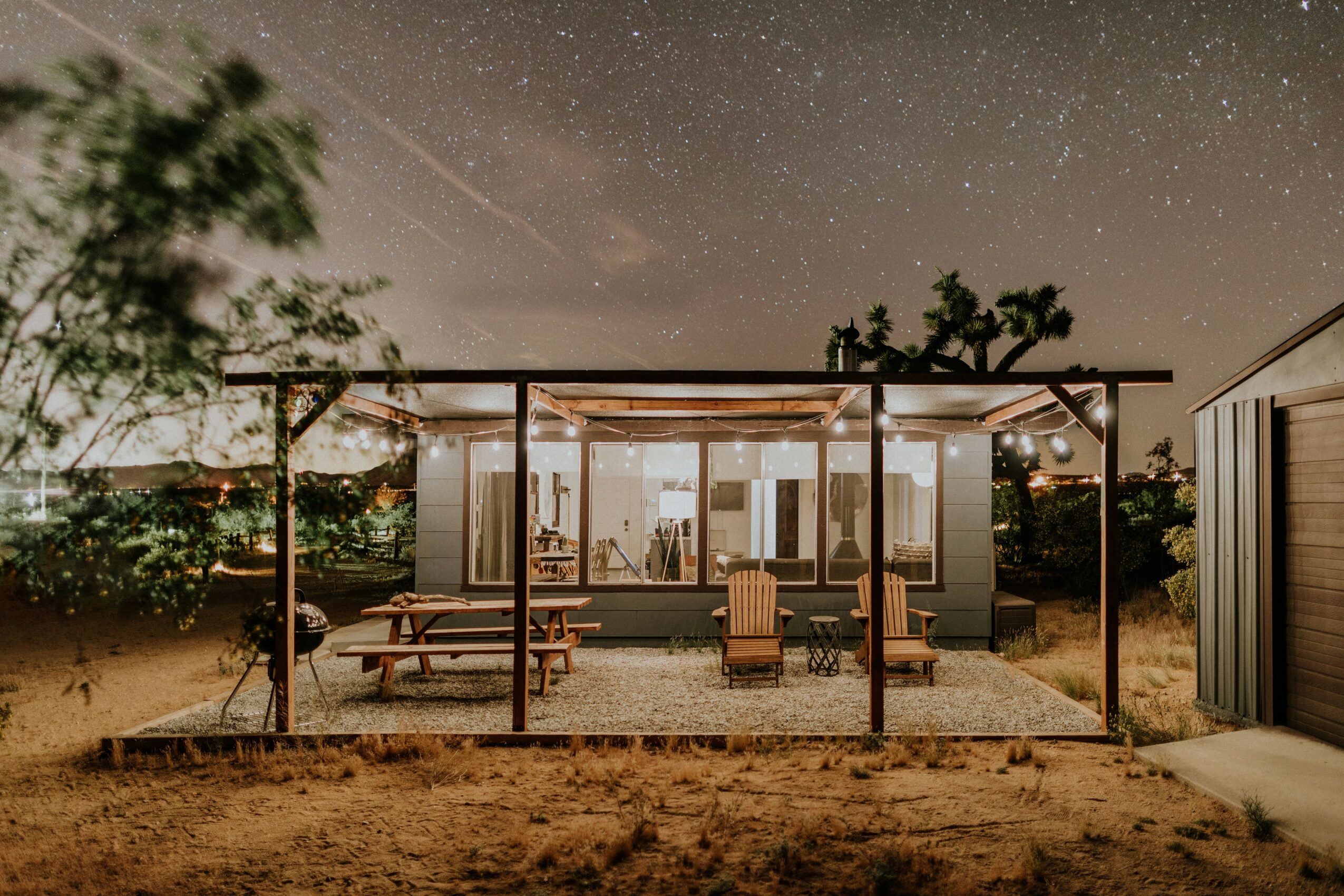Nighttime outdoor patio with seating area, trees, and starry sky, viewed from ground level, with a covered structure and furniture.