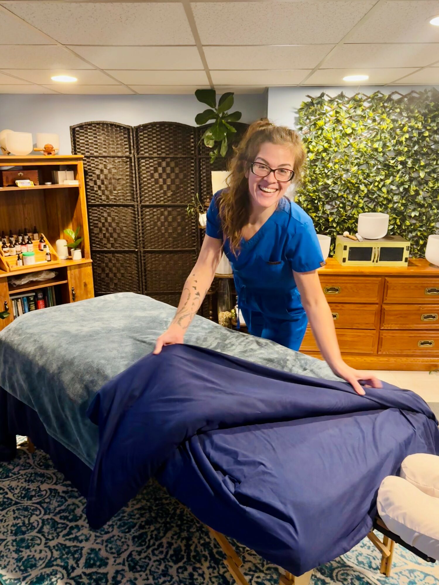 Woman with glasses and blue dress smiling at camera, standing beside massage table in room with bookshelf and greenery.