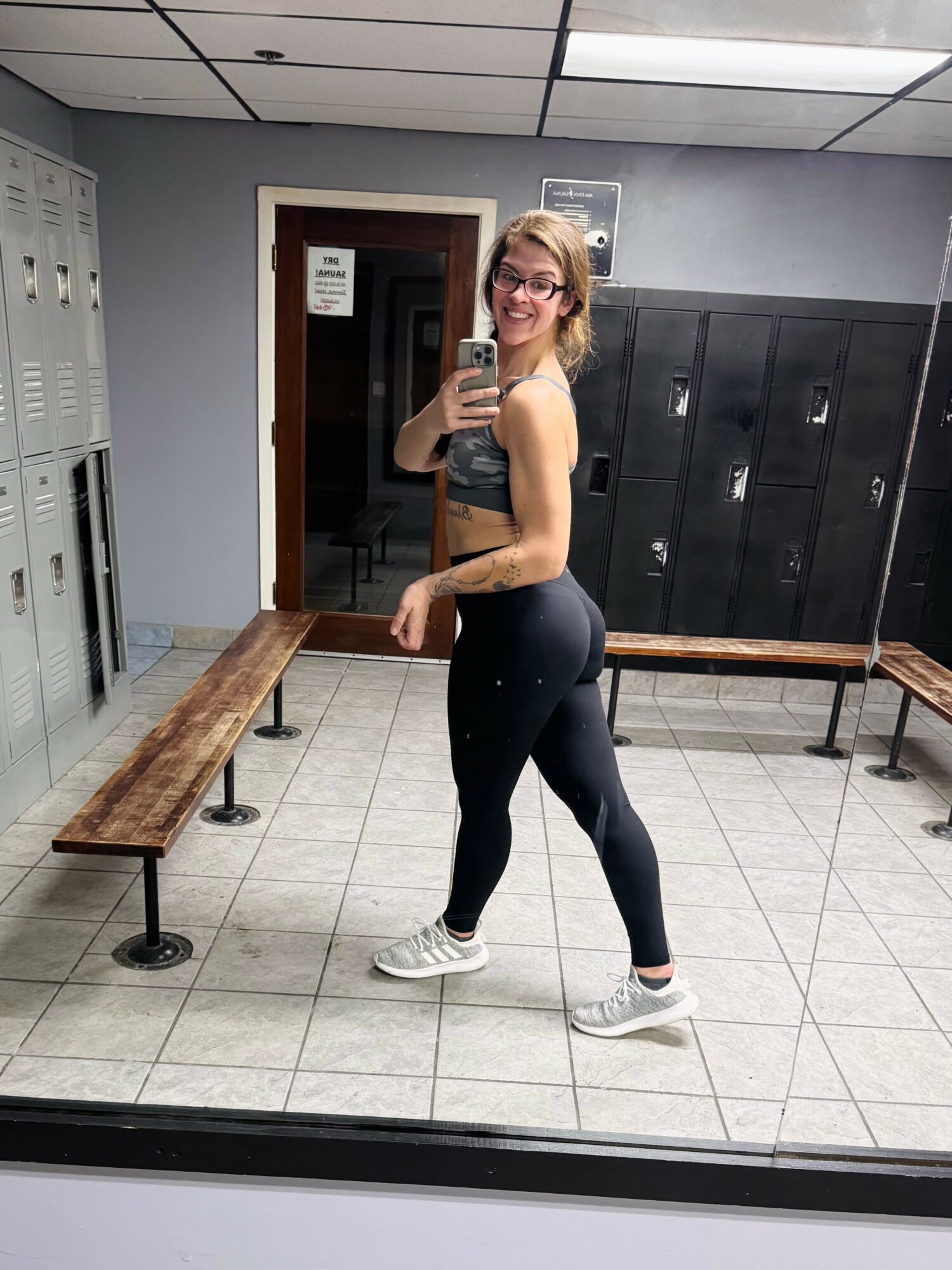 Young woman taking a mirror selfie in a locker room, wearing workout clothes and glasses, smiling, with lockers and benches in background.