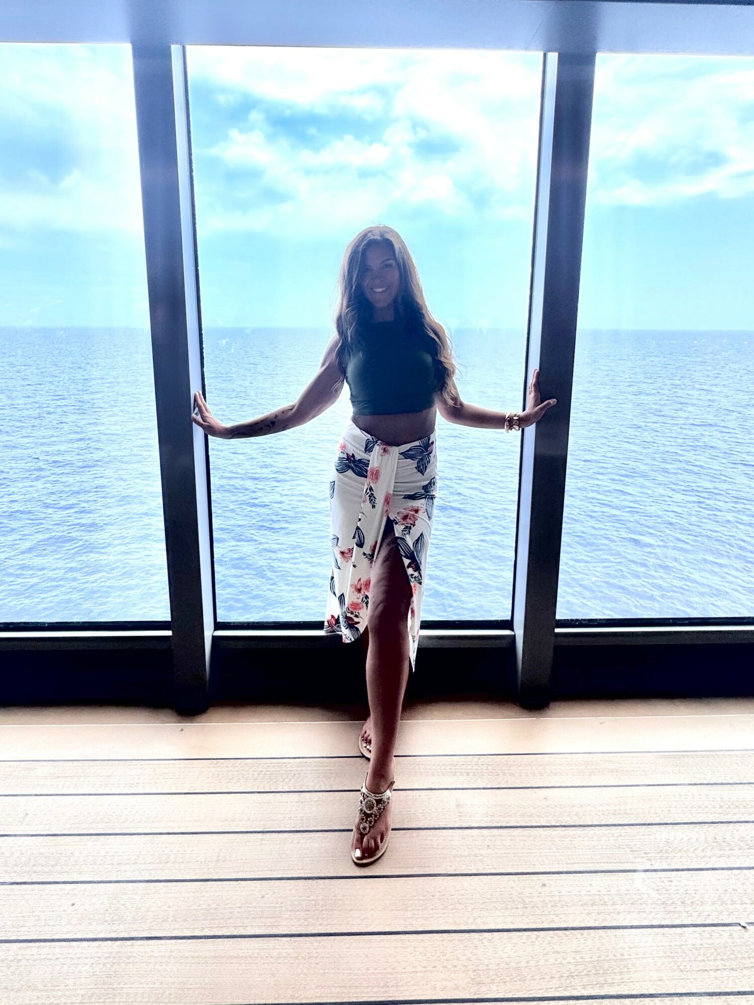 Woman standing in front of large window overlooking ocean, holding sides of window frame, smiling, wearing a dark top and patterned skirt.