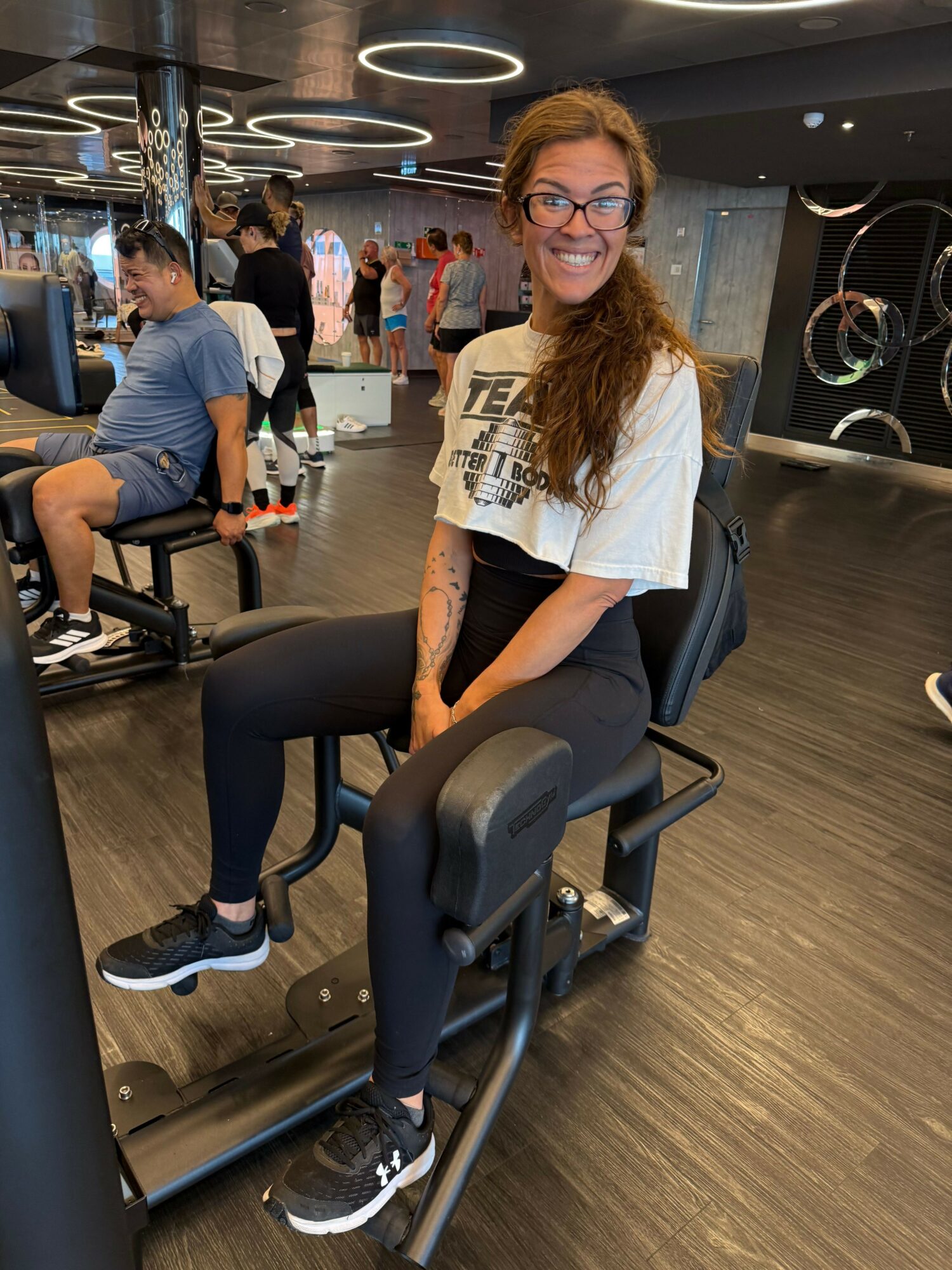 Woman with glasses and curly hair sitting on a fitness machine in a gym, smiling at the camera.
