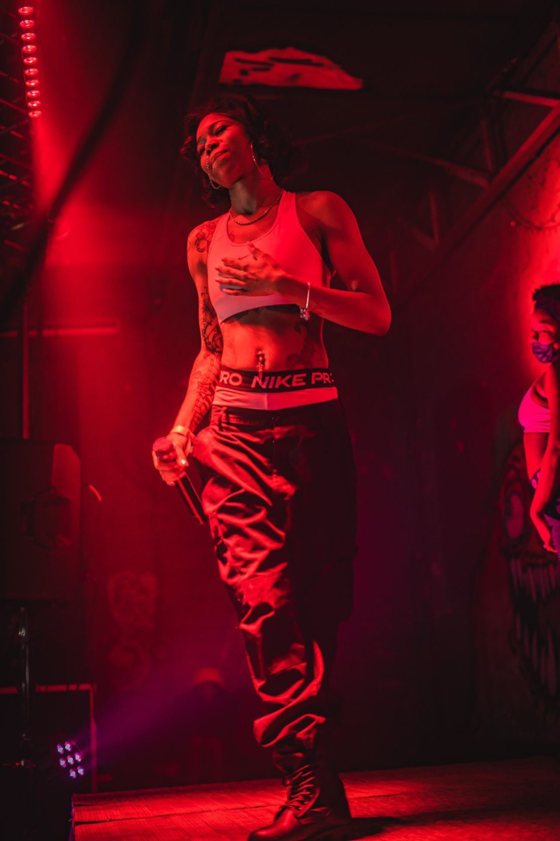 Female dancer on stage with red lighting, wearing a crop top and baggy pants, holding a microphone.