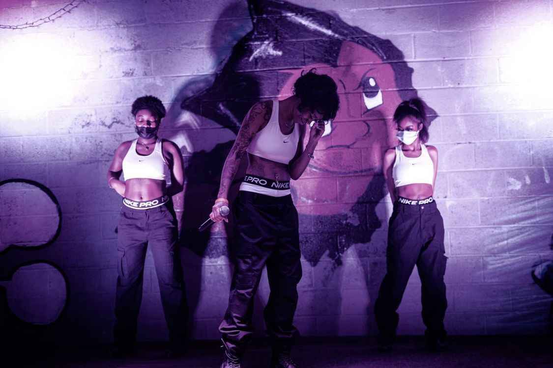Three dancers perform in front of a graffiti wall, wearing masks and casual clothing, with purple lighting.
