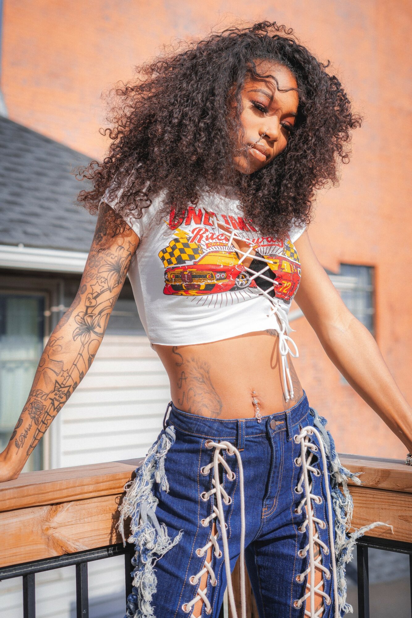 Young woman with curly hair wearing a graphic crop top and lace-up jeans outdoors, leaning on a railing.