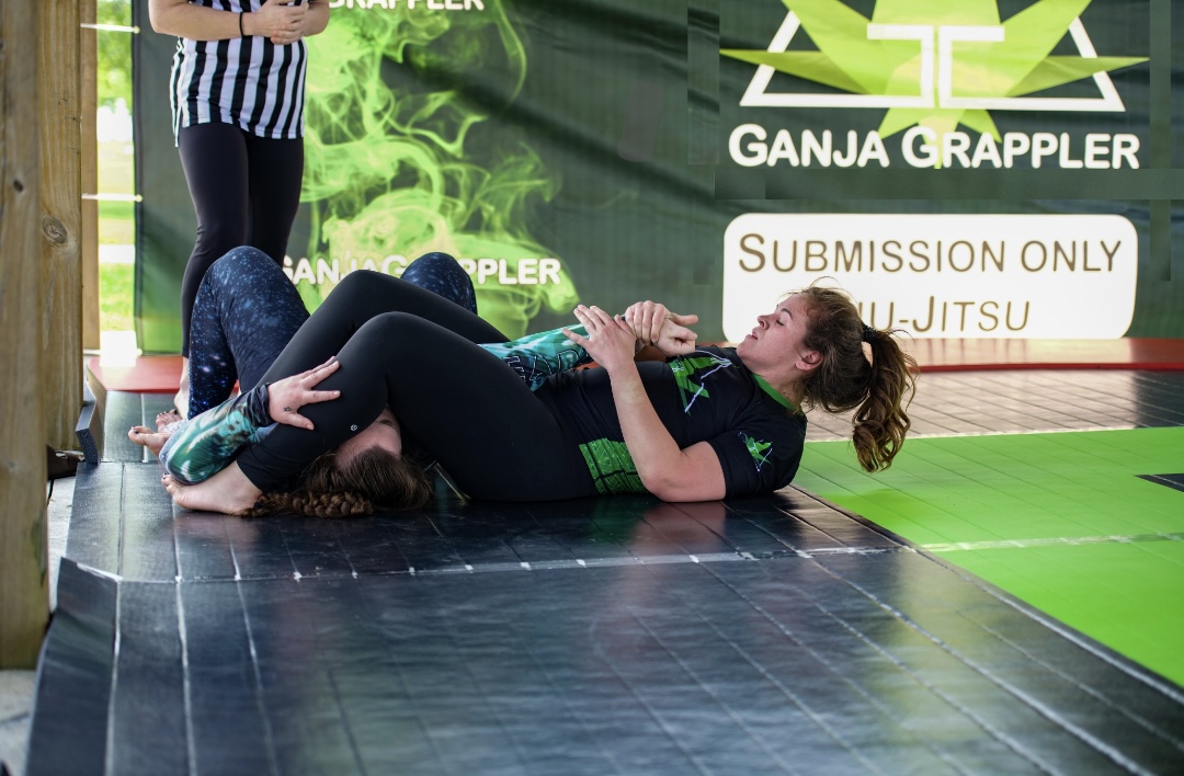 Two women practicing grappling on the floor at a martial arts gym, with a green and black background and a sign reading 'SUBMISSION ONLY' and 'Jiu-Jitsu'.