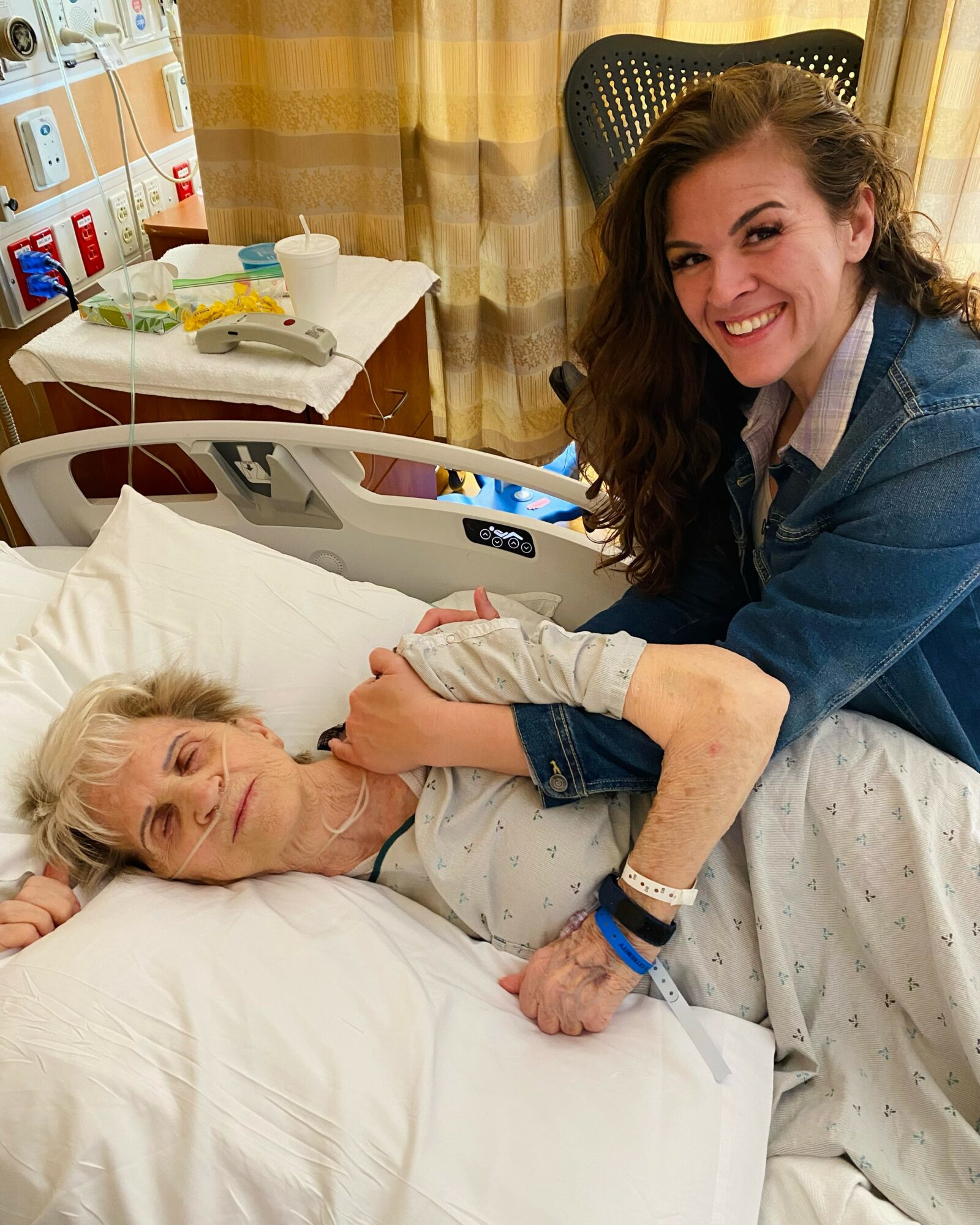 Young woman with long curly hair smiling and holding an elderly woman’s hand in hospital bed.