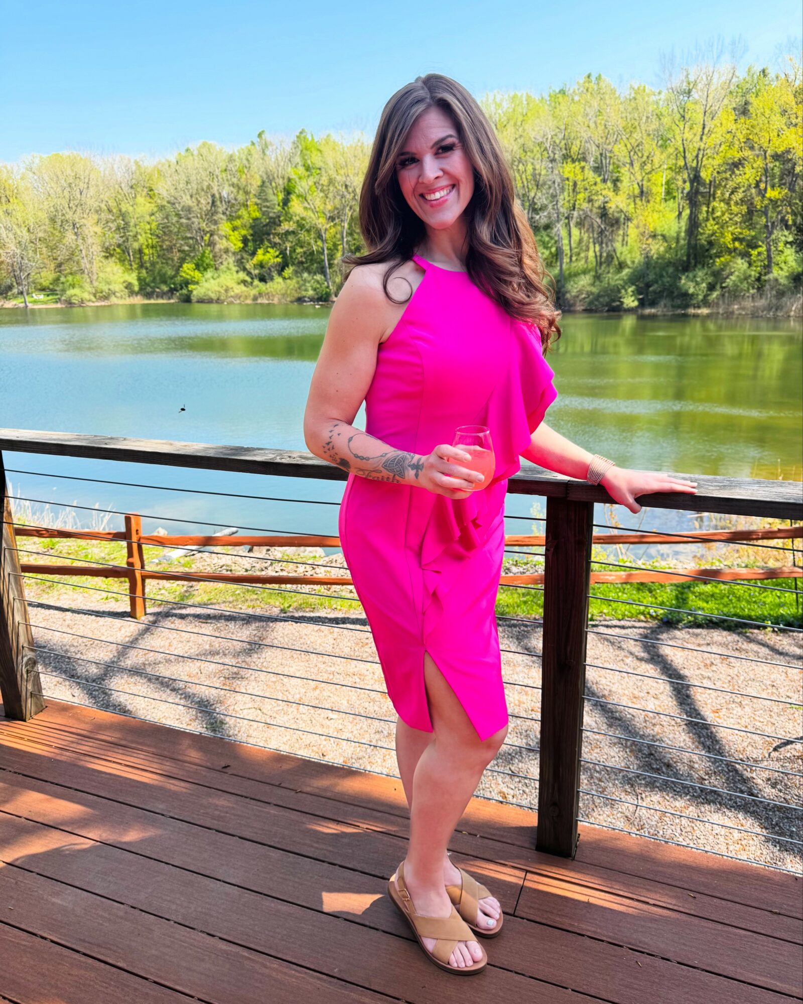Woman in pink dress standing on wooden deck near lake with trees in background, smiling, holding railing.