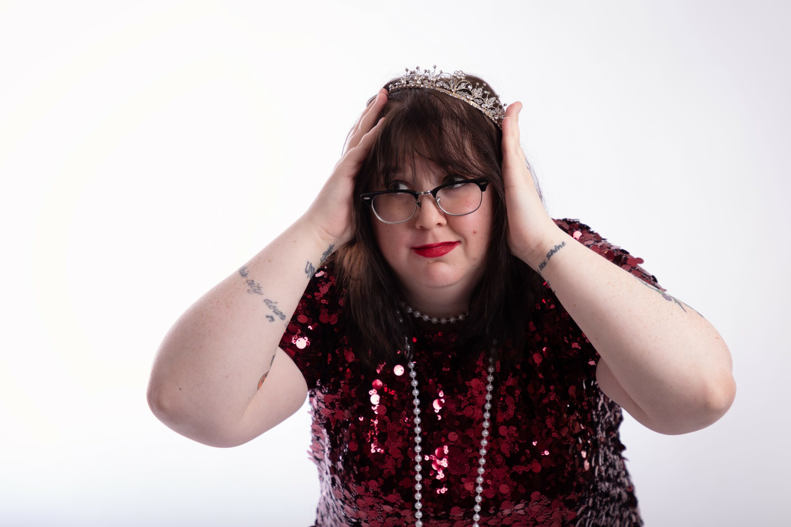Woman with glasses and dark hair wearing a red sequined dress and a tiara, holding her head with both hands.