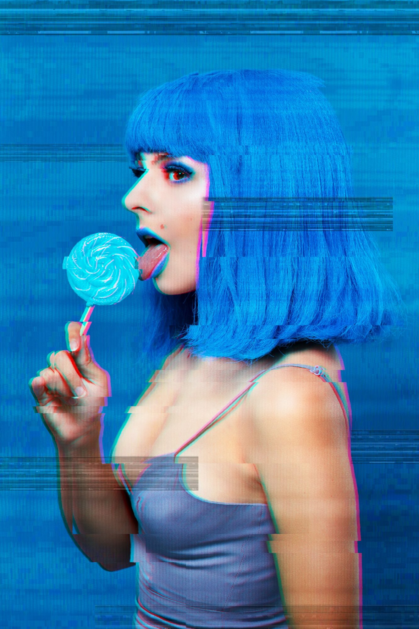 Woman with blue hair licking a blue lollipop, wearing a gray top, against a blue background.