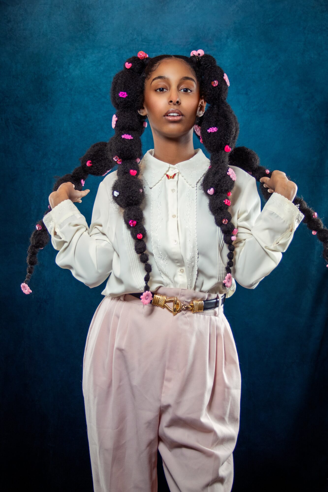 Young woman with styled braids decorated with pink beads, wearing a white blouse and light pink pants, standing against a blue background.