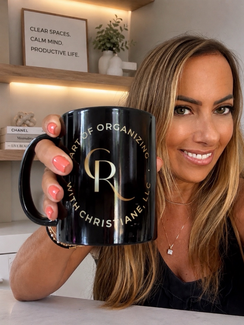 Woman smiling holding a black mug with gold text, in a room with shelves and decor in the background.
