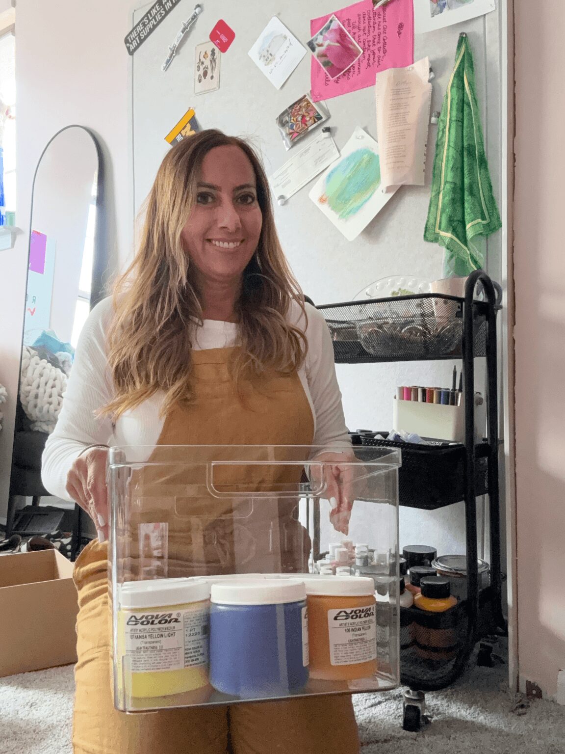 Woman with long hair smiling, standing in a room with a whiteboard, shelves, and various items, holding a clear container.