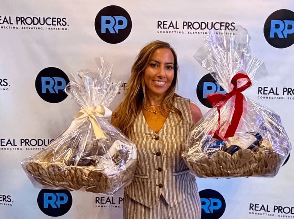 Woman holding two gift baskets wrapped in clear plastic with ribbons, smiling, standing in front of a backdrop with logos.