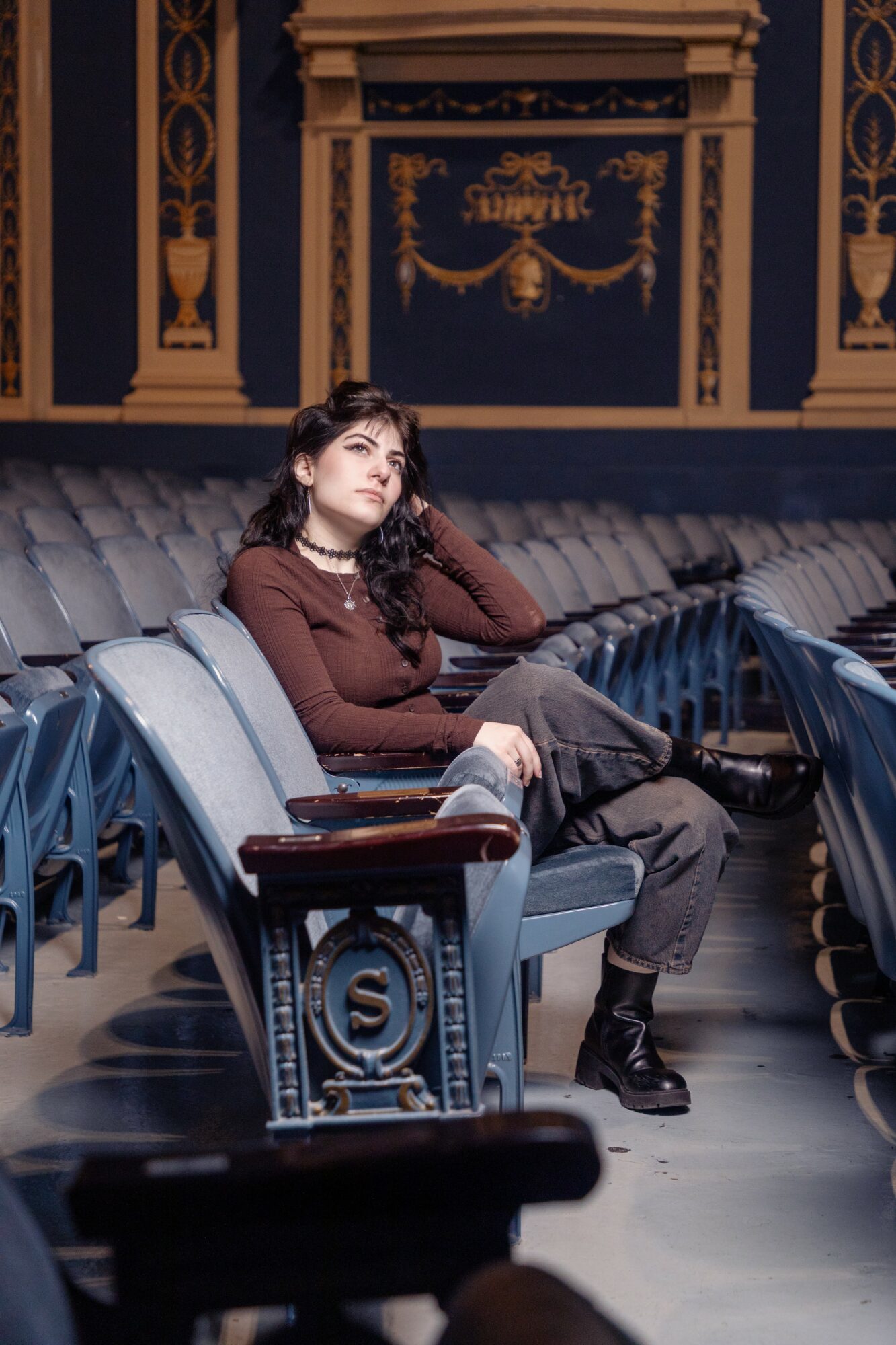 Young woman with curly hair sitting on theater seats, resting her head on her hand, in an ornate theater interior.