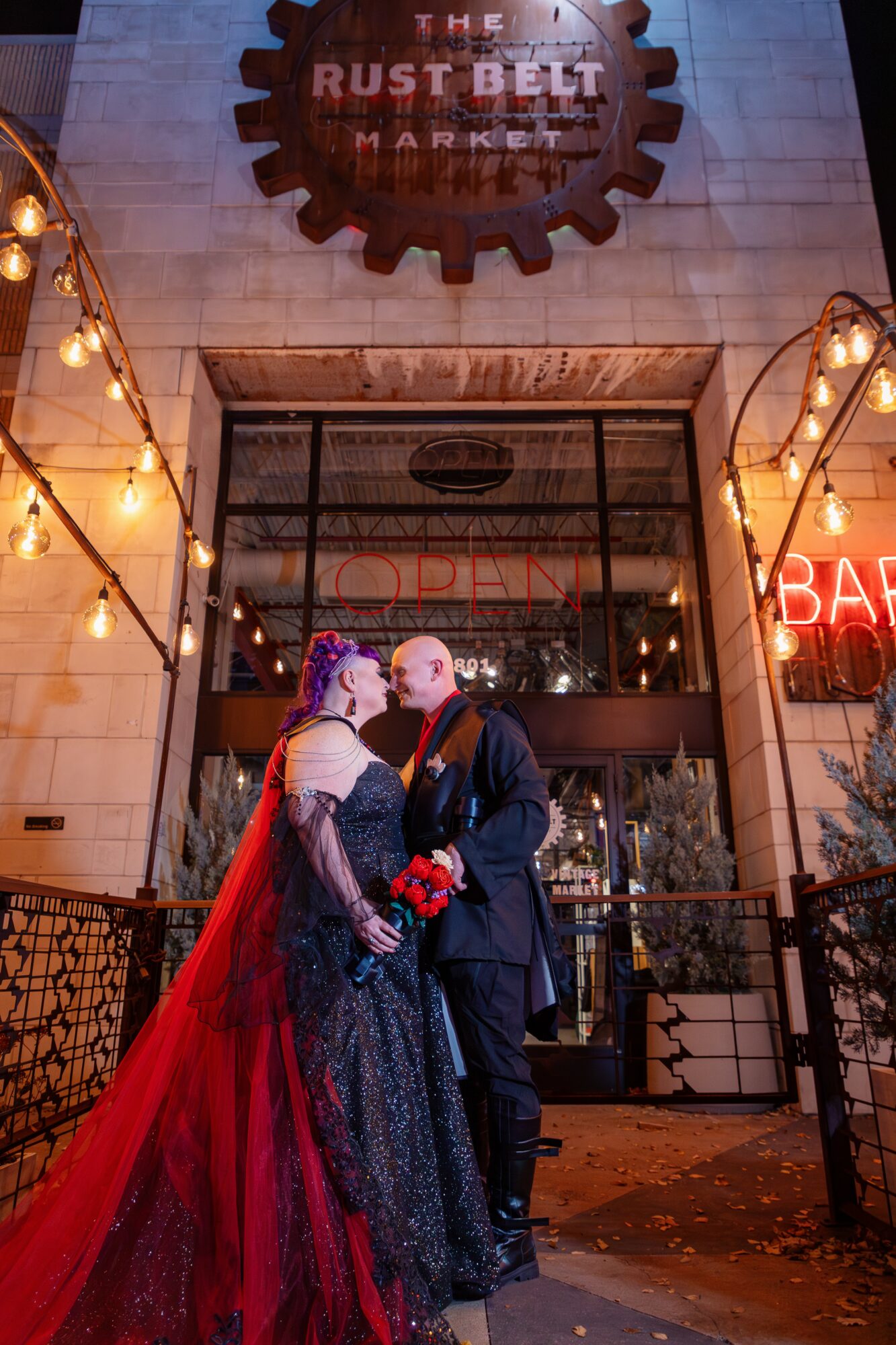 Couple dressed in gothic attire standing close outside Rust Belt Market at night, illuminated by string lights and neon sign.