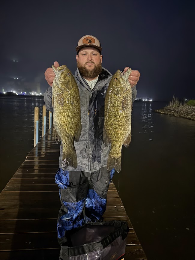 Man holding two large fish on a dock at night, water and city lights in background.