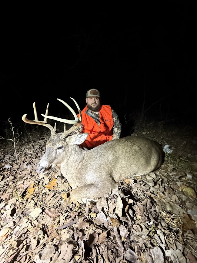 Person in orange jacket and cap kneels behind a large deer with antlers on a rocky ground at night.