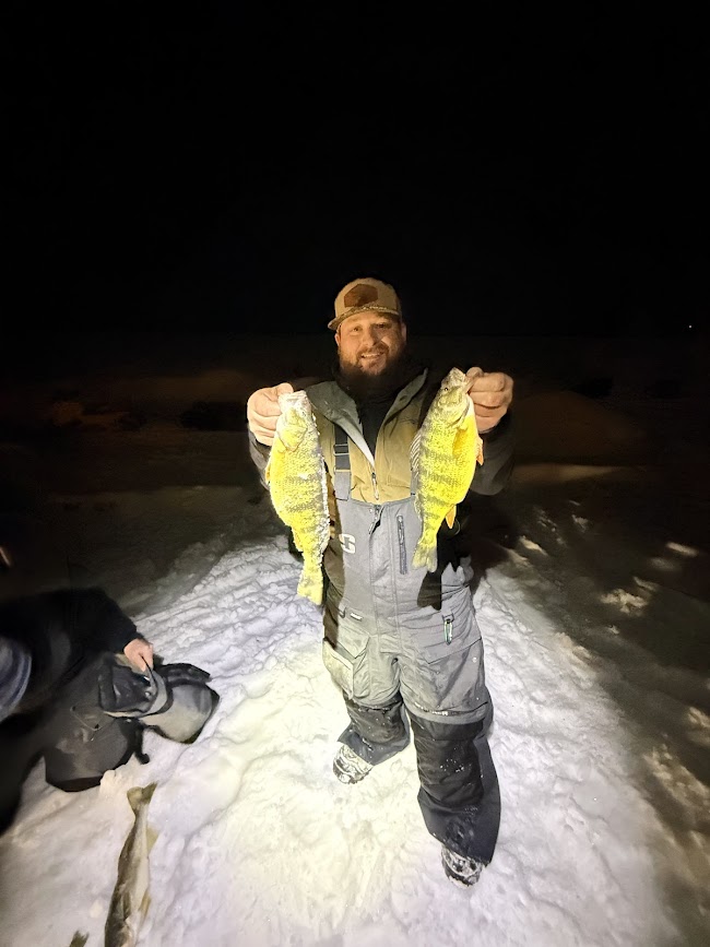 Person holding two yellow fish outdoors at night, snow on ground, person wearing winter clothing, dark background.