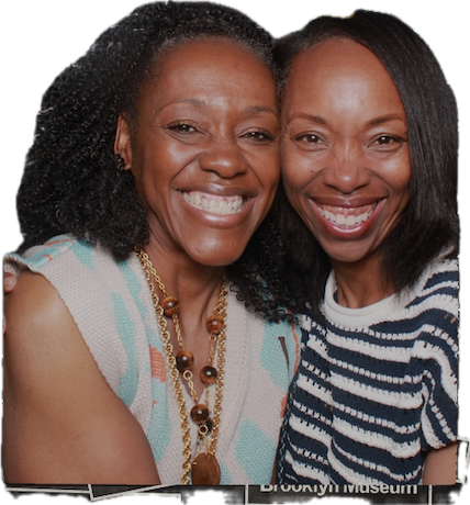 Two women smiling closely, one with curly hair and the other with straight hair, wearing striped and patterned tops.