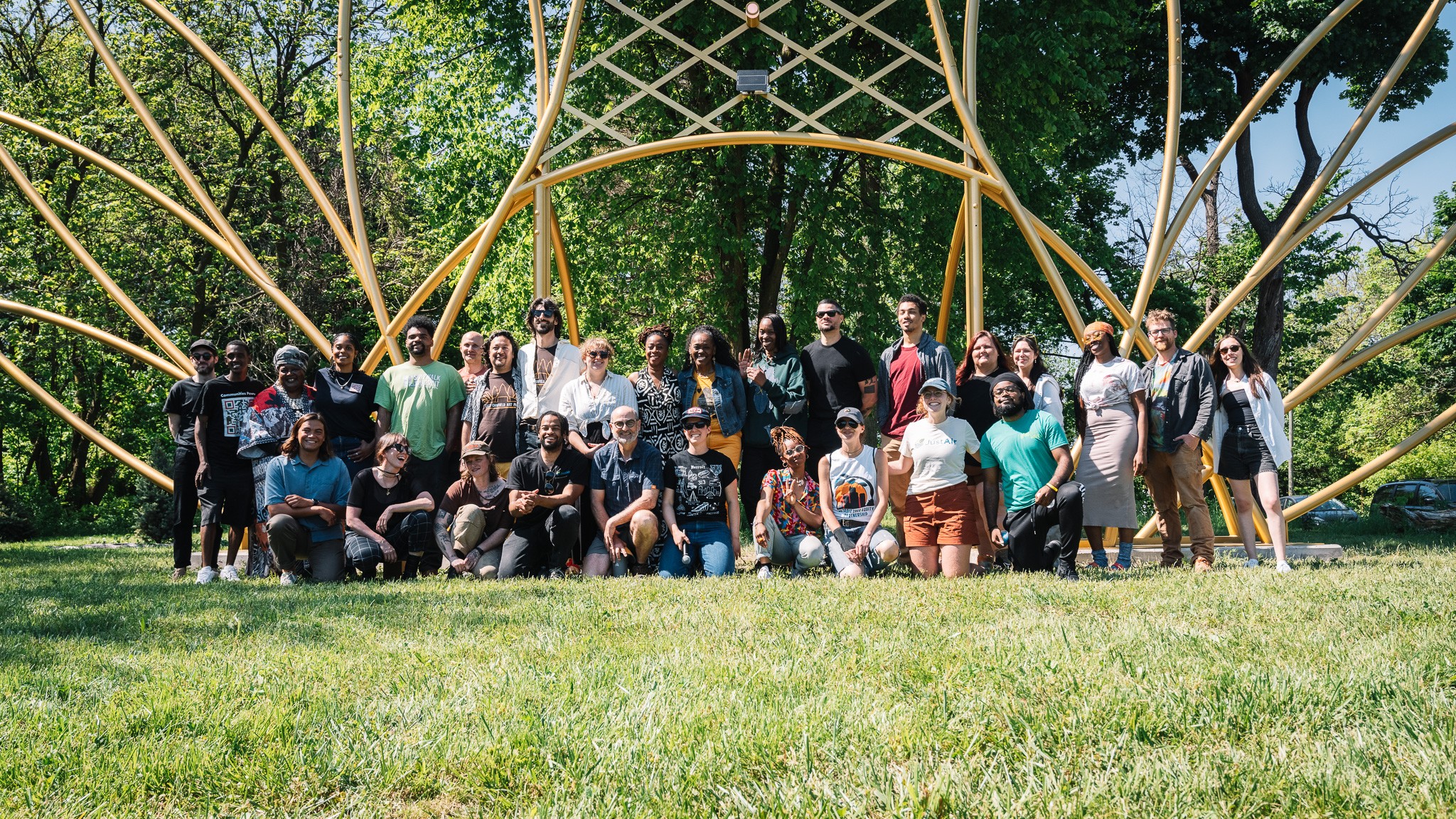 Group of people outdoors in front of a large, abstract metal sculpture, with trees and grass in the background.