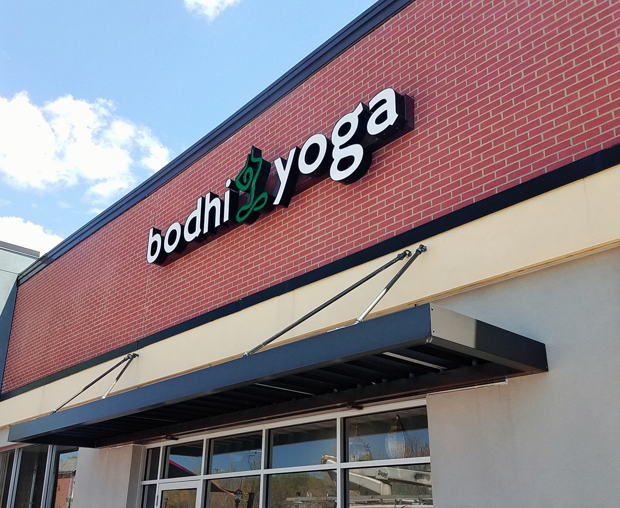 Storefront with sign reading 'bodhi yoga' on a brick building, under a black awning, with a blue sky background.