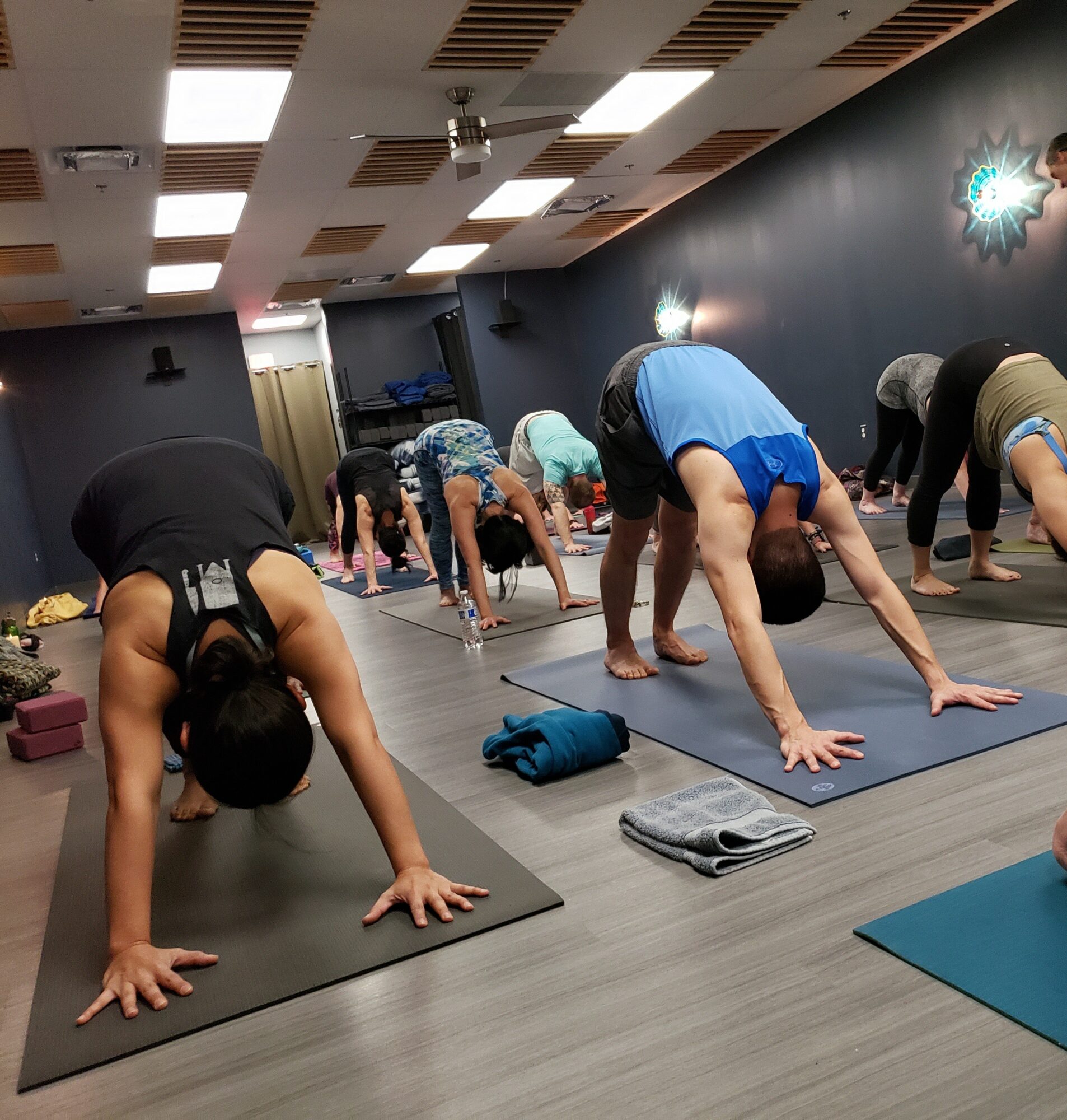 People participating in a yoga class in a studio, bending forward on mats, with a dark wall and ceiling lights.