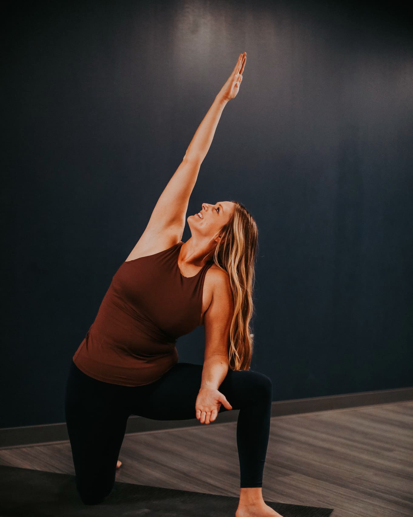 Woman in yoga pose with one arm raised, kneeling on one knee, against a dark wall background.