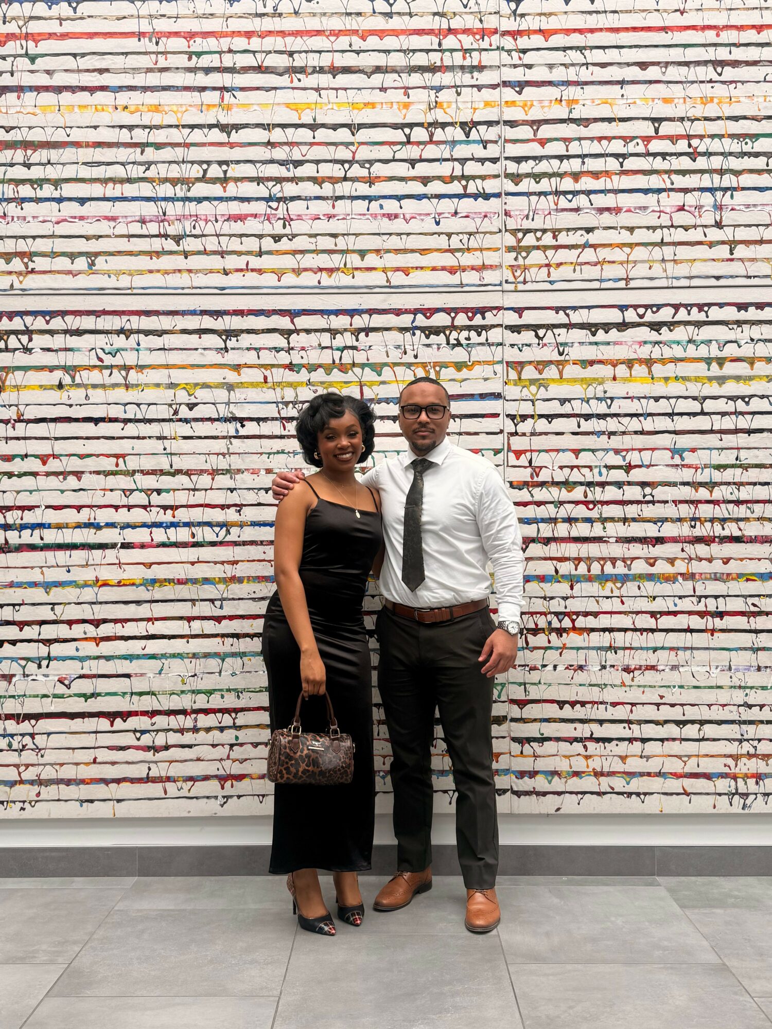 Two people standing in front of a colorful, vertical striped wall, one holding a purse, both facing the camera.