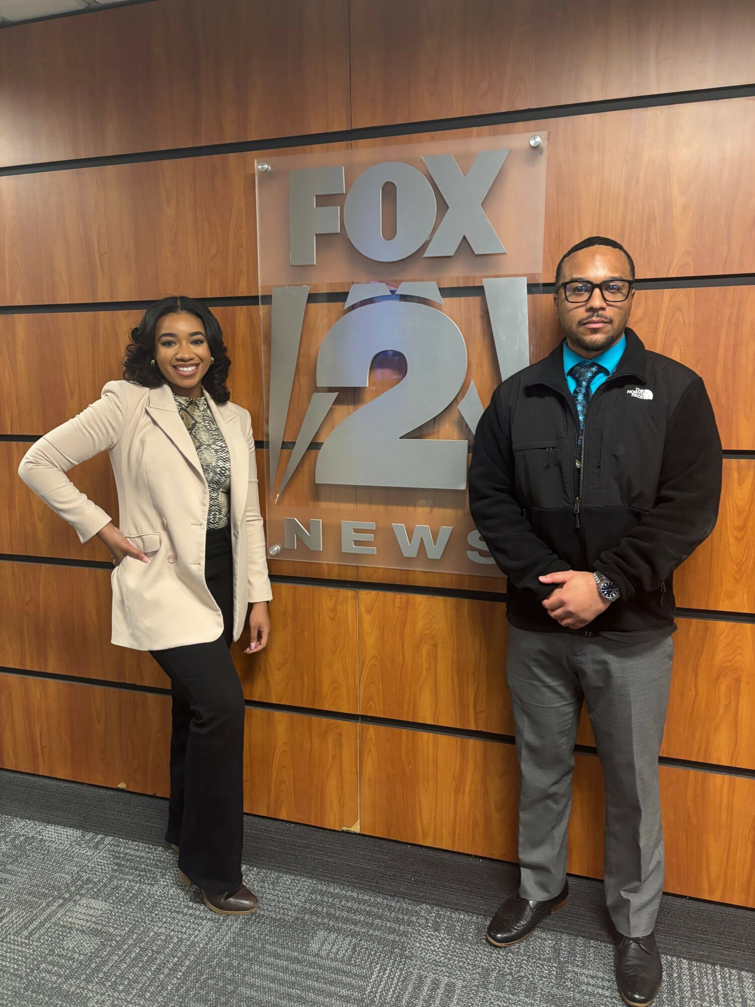 Two people standing in front of a FOX 2 sign, one in a black jacket and the other in a white coat, both smiling.