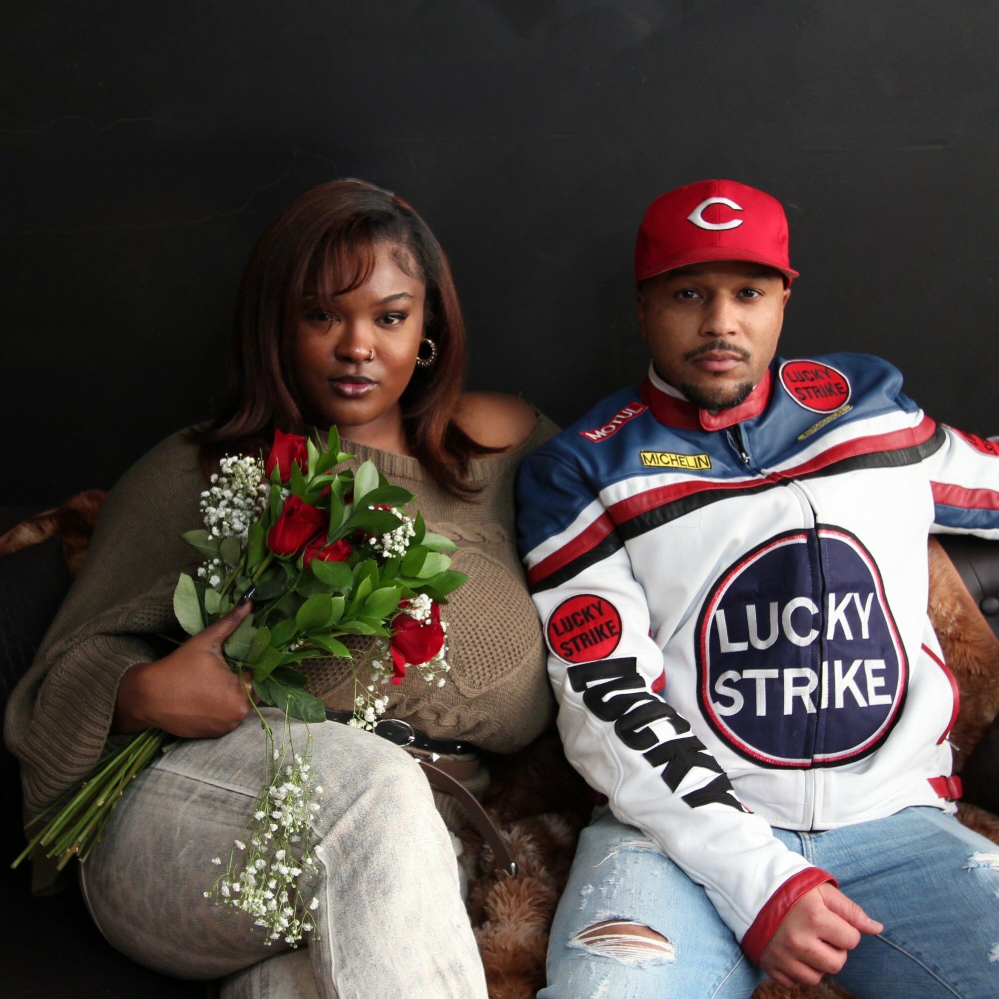 A woman and a man sit together against a dark background, holding a bouquet of flowers, both looking at the camera.