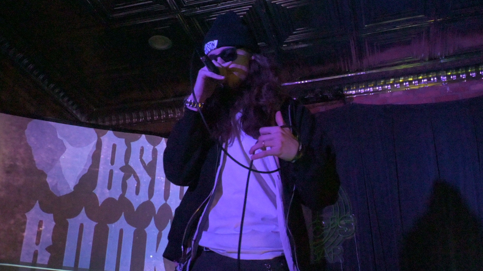 Person wearing a cap and jacket holding a microphone on stage with a dark background.