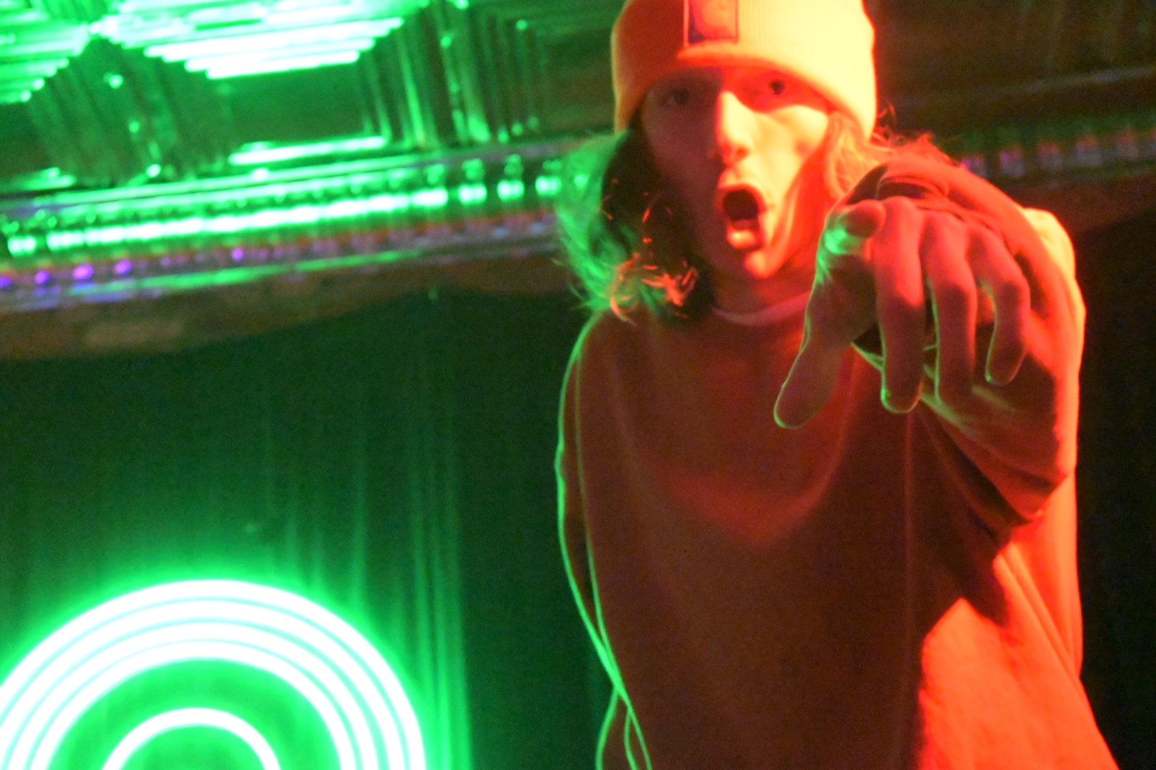 Person wearing a beanie pointing towards the camera with a surprised expression, illuminated by green and red lights.